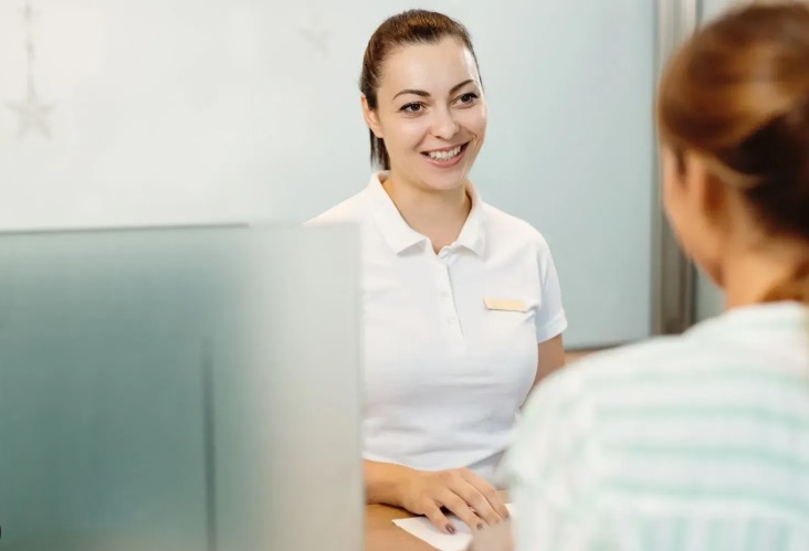 Imagen personal, protocolo y entorno físico en clínicas estéticas: cómo la recepcionista proyecta la marca del centro 1 Recepcionista de clínica atendiendo con una sonrisa a una paciente en el mostrador